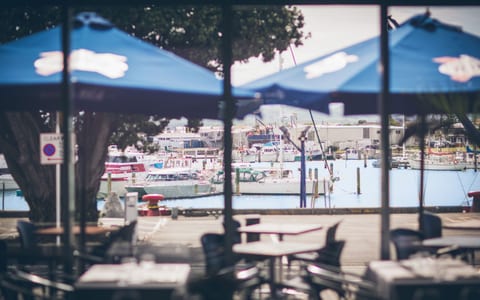 Outdoor terrace with blue parasols overlooking a marina with moored vessels