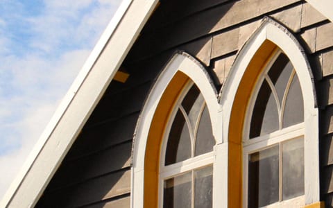 Grey weatherboard gable end with arched Gothic-style windows and yellow surrounds against blue sky