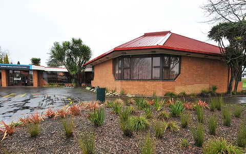 Brick community hall with red metal roof and planting beds on a wet day
