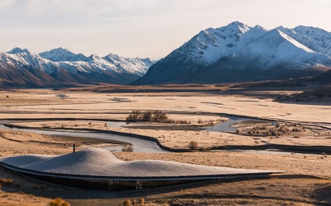 Low-profile curved building on a flat river plain with snow-capped mountains behind