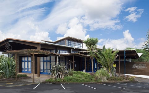 Low-rise timber and dark-stained building with pergola canopy and planted entry, car park