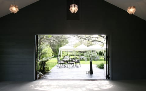 A dark barn-style interior with three crystal chandeliers and open bifold doors onto a deck, garden and white marquee