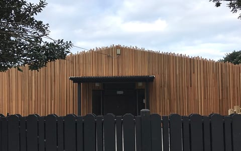 Low single-storey timber-clad building with vertical battens and jagged roofline, dark fence in foreground