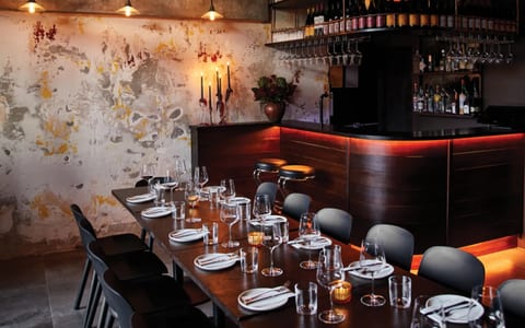 Dark dining room with distressed plaster wall, warm-lit timber bar, pendant lights, and set dining table