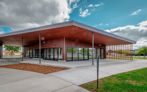 Low flat-roofed timber building with a wide canopy, glass frontage, and paved surrounds on open grounds