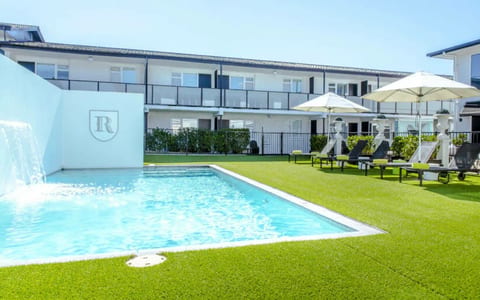 White two-storey hotel building with pool, sun loungers, umbrellas, and manicured lawn