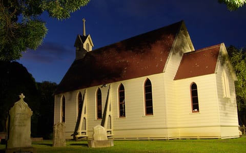 Small white timber church illuminated at night with historic graves in foreground