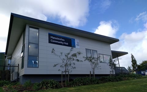 Single-storey community hub building with flat roof, signage board, and garden border