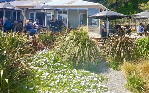 An outdoor seating area with dark umbrellas and picnic tables among native flax beside the hotel