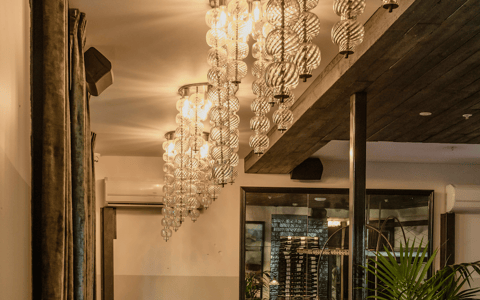 Warm dining room with globe chandeliers, timber ceiling, banquette seating, and wine display beyond glass partition