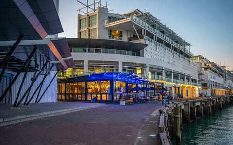 Waterfront bar and dining pavilion with blue neon canopy, dusk, harbour backdrop