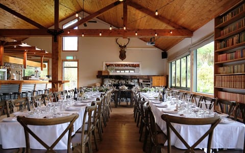 High-stud timber-framed reception hall with long banquet tables, a stone fireplace and string lights