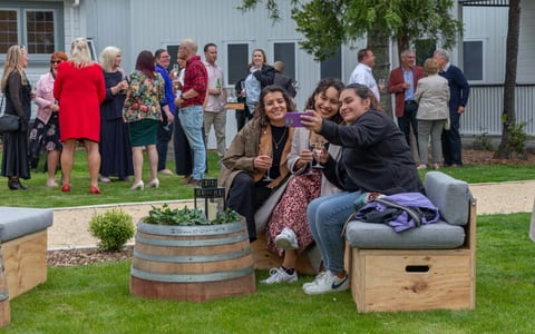 Guests gathered on a lawn beside a white weatherboard building with outdoor barrel furniture