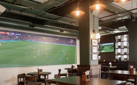 Dark green bar interior with large screen, plaid carpet, and high wooden tables