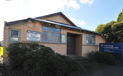 Timber-clad community hall with dark door, signage and overgrown shrubs