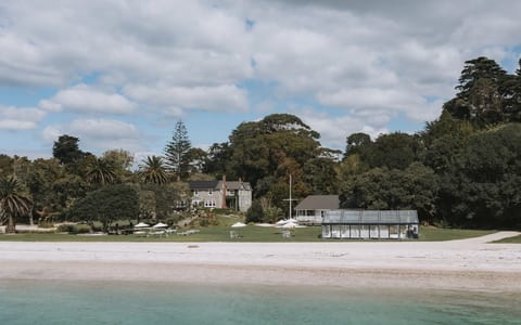 Stone homestead and glass marquee on lawn beside sandy beach with trees behind, viewed from water