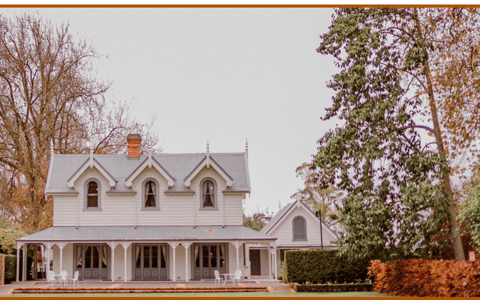 White Victorian-era homestead with steep roof, dormer windows and wrap-around veranda in formal hedged gardens with autumn trees