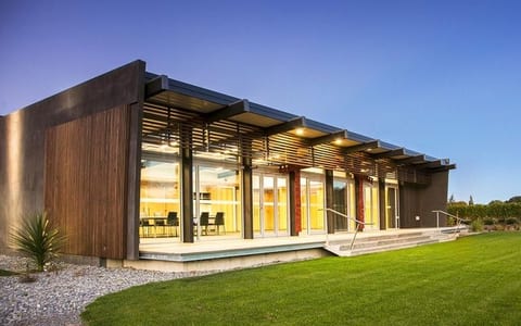 Low dark-clad building with timber louvred pergola, lit interior visible through full-height glazing, lawn foreground at dusk