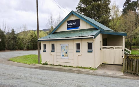 Small cream weatherboard hall with green metal roof beside a gravel road