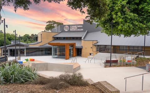 Brick and metal-roof building with timber-framed glazed entry, concrete forecourt, gardens at dusk