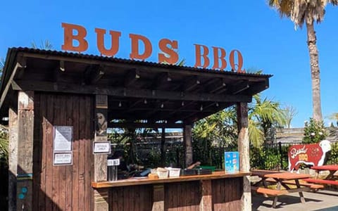 Outdoor rustic bar kiosk with Buds BBQ signage and picnic seating