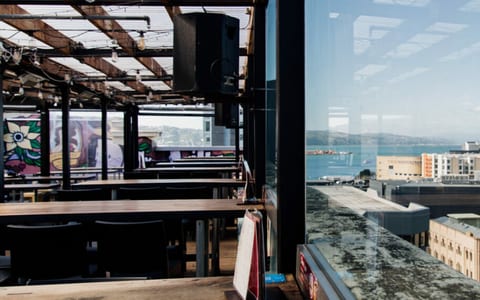 Open rooftop bar interior by day with exposed steel beams, long wooden benches, and large window overlooking city