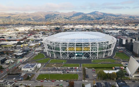 Large oval stadium with white lattice steel frame and translucent roof, aerial view in an urban setting with hills behind