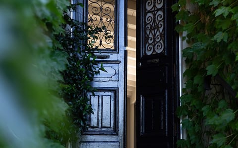 Dark iron-wrought entrance doors with ornate scrollwork panels, ajar to reveal lit interior, stone steps and climbing plants