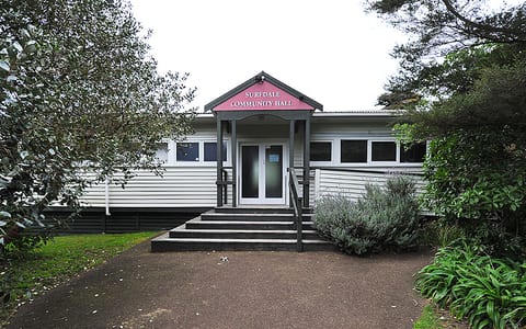 White weatherboard hall with entry steps and pink fascia sign, surrounded by planting