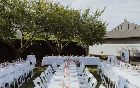 Outdoor reception with white linen tables, white folding chairs, festoon lights and mature trees
