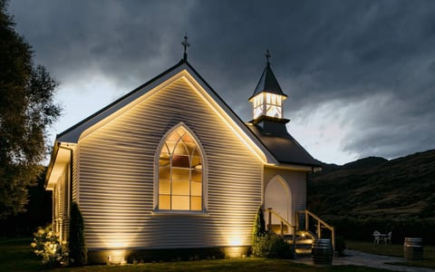 White timber church exterior lit at night with glowing arched window and dark hill beyond