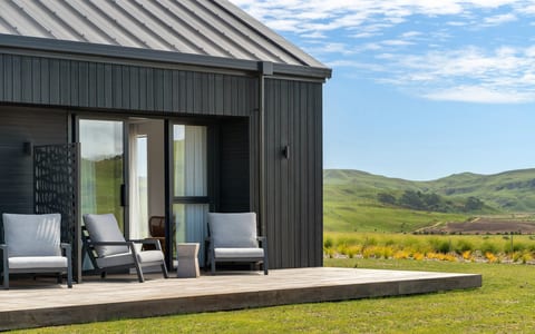 Dark vertical-clad building with timber deck, two outdoor chairs, and green hillside view