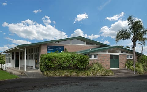Low-set brick hall with green metal roof, covered verandah, and a palm tree alongside