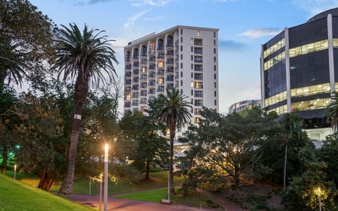 Tall white hotel tower with arched windows, seen from a park with tall trees at dusk
