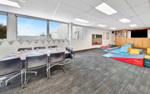 Carpeted activity room with round table, chairs and foam play equipment beside windows