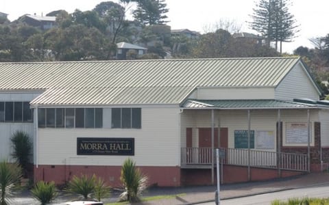 Low-pitched cream and red-brick community hall exterior with green corrugated roof, hillside behind