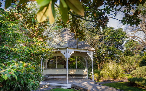 White wooden gazebo with shingled roof set among dense mature trees and garden
