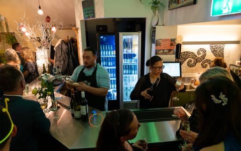 Bar service area with illuminated drinks fridge and bartender, decorated interior space with guests in background