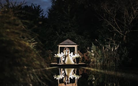 Illuminated gazebo on a jetty reflected in a still pond at night, surrounded by trees