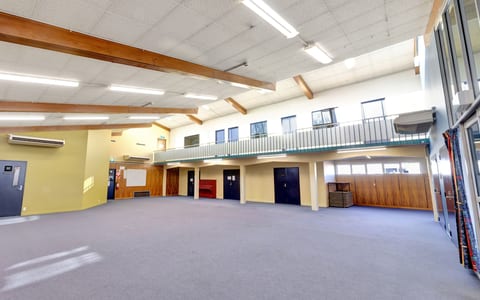 Large cleared hall with grey carpet, exposed timber roof beams, mezzanine balcony, and high windows