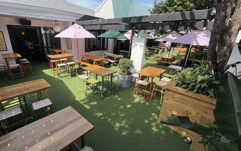Open-air courtyard with timber picnic tables, coloured stools, market umbrellas, and turf underfoot