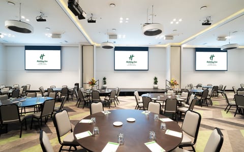 Large carpeted function room with round tables, three projection screens, and recessed ceiling lighting