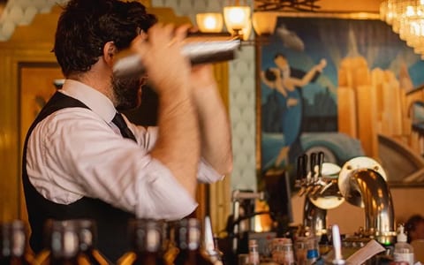 Bartender shaking cocktail behind bar with art deco mural and beer taps