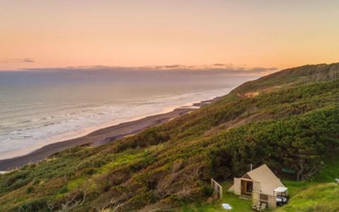 Canvas tent structure on grassed hillside at sunset, overlooking black-sand beach and ocean