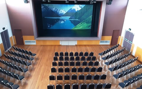 Auditorium with rows of black chairs facing a large projection screen on a raised platform, timber floor