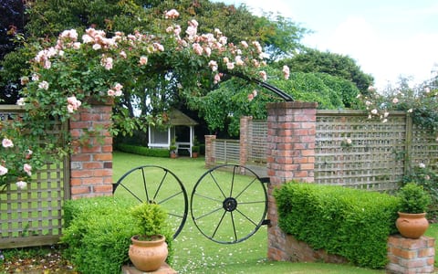 Brick garden gateway with climbing roses and black iron wagon wheels, gazebo beyond on lawn
