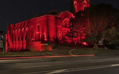 Red brick church with castellated tower, illuminated in red at night, viewed from across the road