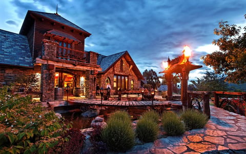 Stone and timber estate building at dusk with lit torch flares, pond, and garden terrace
