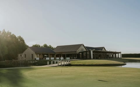 Timber and steel clubhouse beside a putting green with pond and trees, low afternoon light
