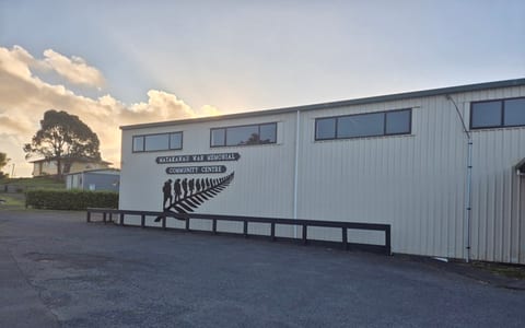 White corrugated-iron hall exterior with fern silhouette mural, car park in foreground